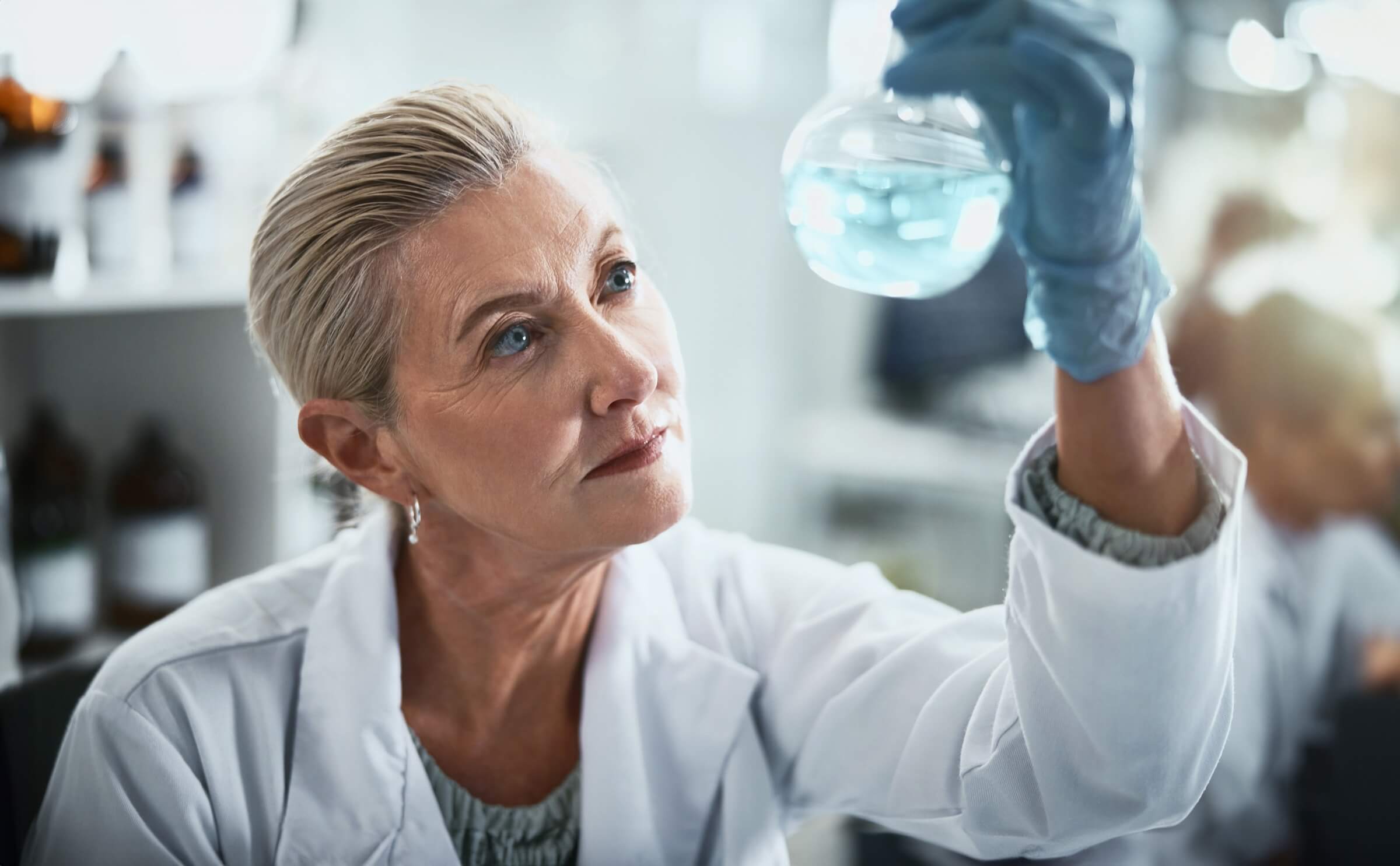 Naukowiec w kitlu laboratoryjnym i niebieskich rękawiczkach trzyma kolbę wypełnioną niebieskim płynem, intensywnie ją badając.