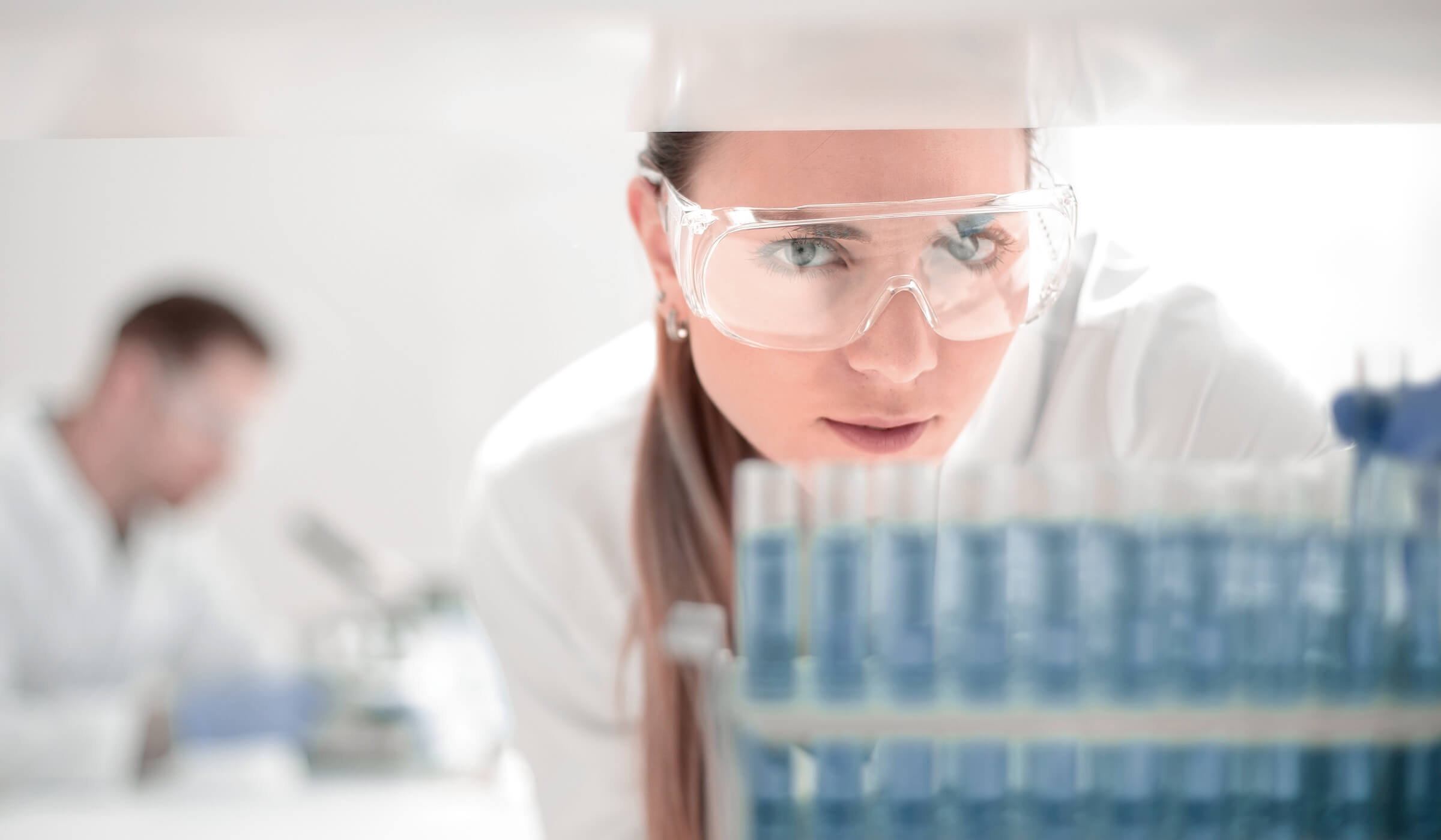 Kobieta w okularach ochronnych i białym kitlu, pochylona nad probówkami z niebieską cieczą w laboratorium.