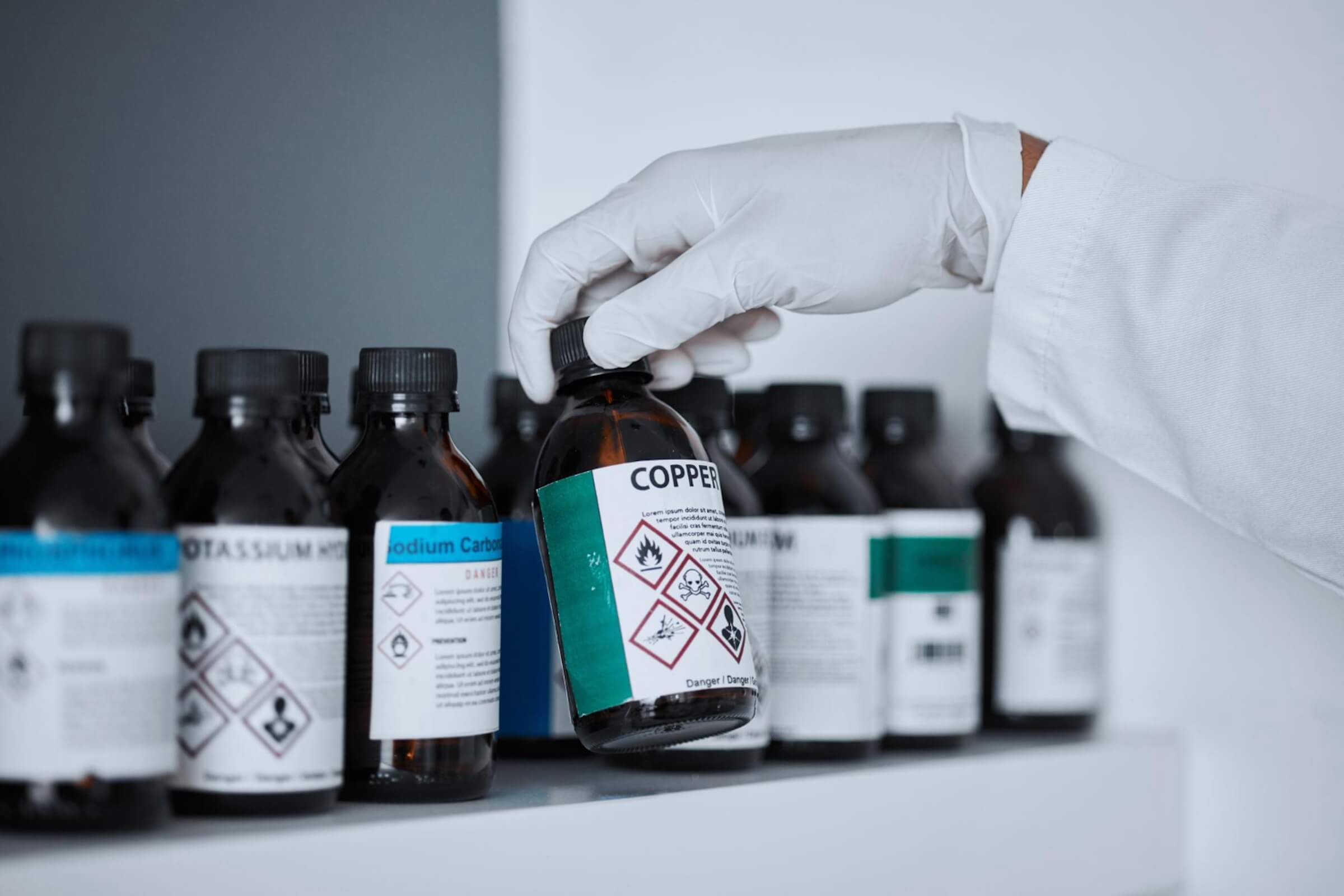 Zbliżenie osoby w białym kitlu i rękawiczkach sięgającej po brązową butelkę z etykietą "COPPER" z półki pełnej chemikaliów